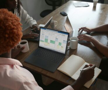Three people in a meeting at a table discussing schedule on their Microsoft laptop